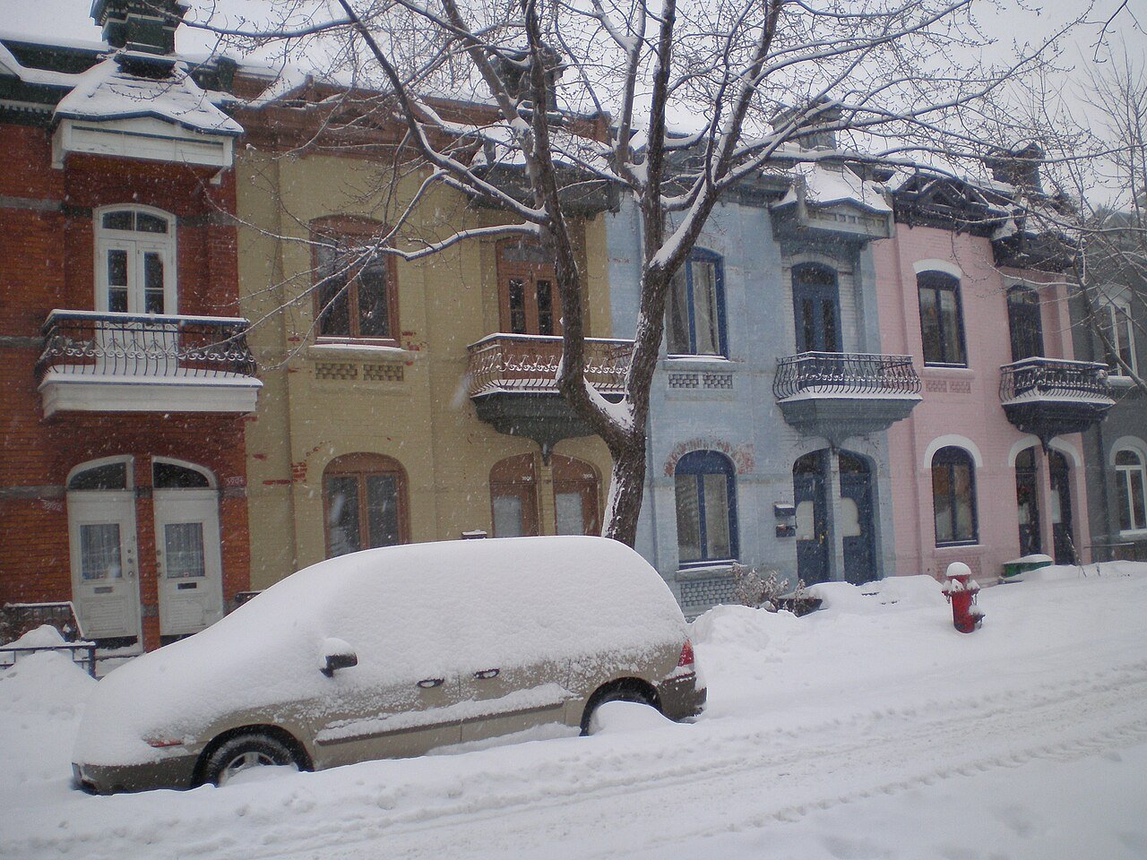 下雪天，Plateau-Mont-Royal 一排红、黄、蓝、粉色的老式联排楼，路边停着一辆被雪盖住的车
